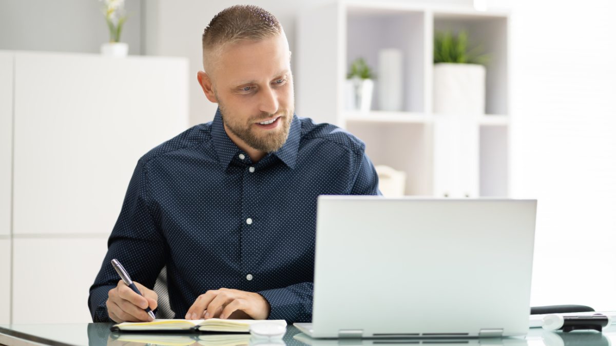 A man wearing a dark blue collared shirt with white dots sits at a reflective desk while looking down at a laptop computer and using a pen to write in a notebook. White shelves in the background hold plants in white pots.