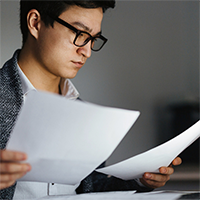 A man wearing glasses sits at a desk, holding a sheet of paper in each hand.