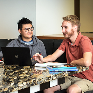 Two male University of Utah students sit at a study space in Marriott Library. The student on the left is wearing glasses and a gray hoodie. The student on the right is wearing beige shorts and a red polo shirt and is pointing at a laptop computer. Image courtesy of the University of Utah.