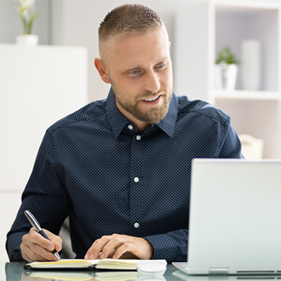 A man wearing a dark blue collared shirt with white dots sits at a reflective desk while looking down at a laptop computer and using a pen to write in a notebook. White shelves in the background hold plants in white pots.